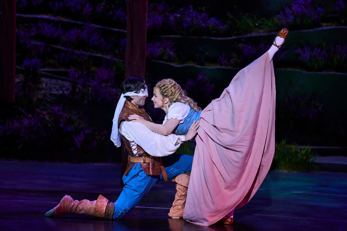 A man kneels blindfolded as a woman leans on him with one leg high in the air.