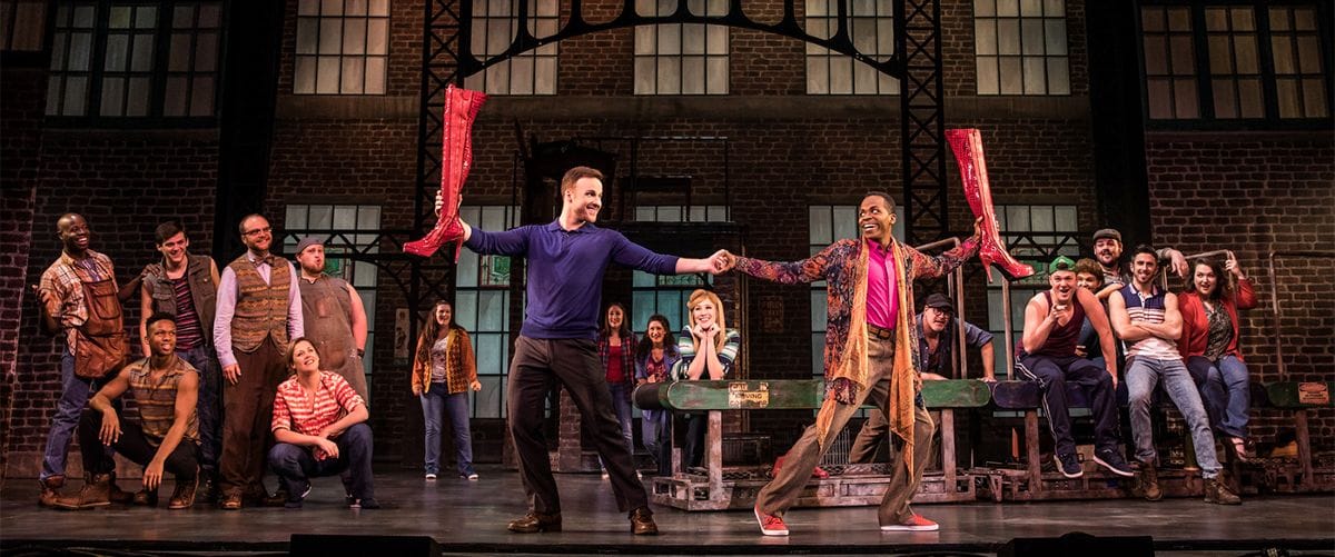 In front of an old industrial building, a group of people watch as two men dance, each holding a red, heeled boot.