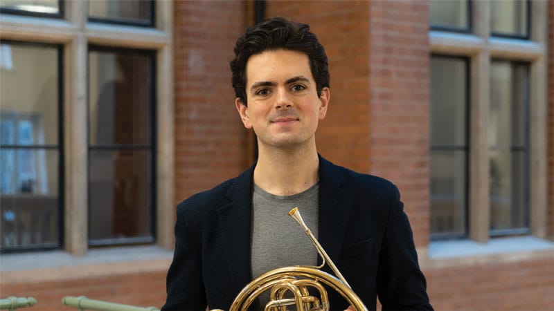 Diego Incertis Sánchez with his french horn in front of a brick-and-windows backdrop.