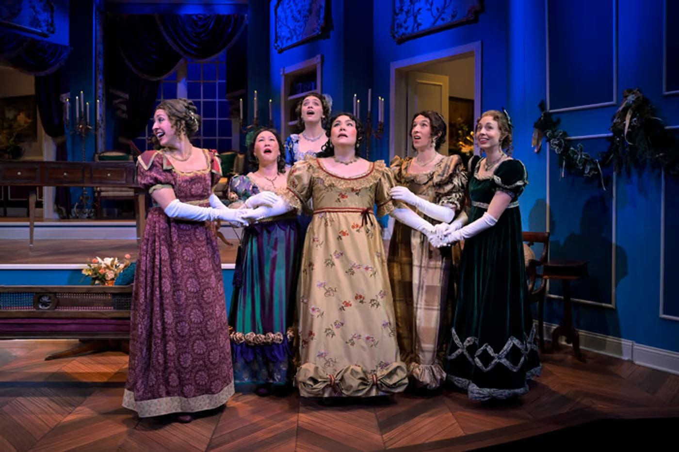 A group of women stand together looking up and excited. They wear regency era dresses in a large house.