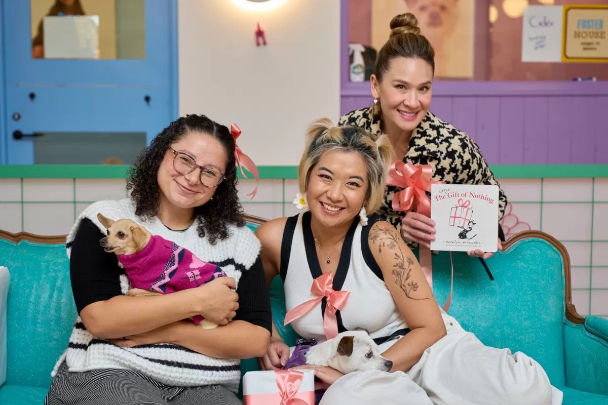Three women sit on a couch holding dogs and gifts, smiling at the camera.