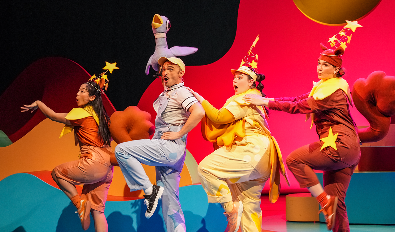 Four actors wear brightly colored outfits with stars on their hats. One is in blue and holding a puppet pigeon.