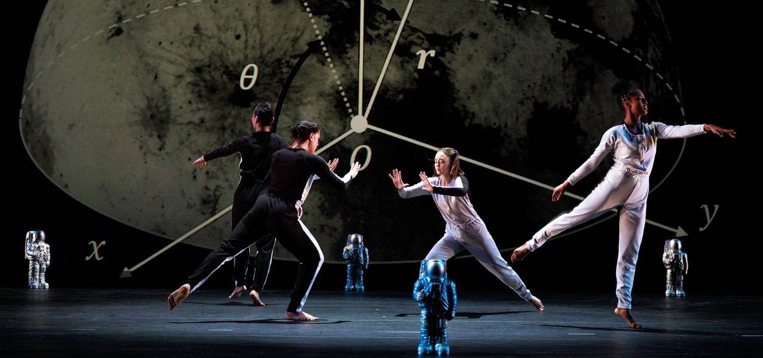 There is a large moon in the background with small astronaut statues. Four dancers are in the forefront dancing.