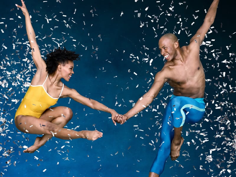 A man and woman dancer are leaped in the air, holding hands with confetti around them.