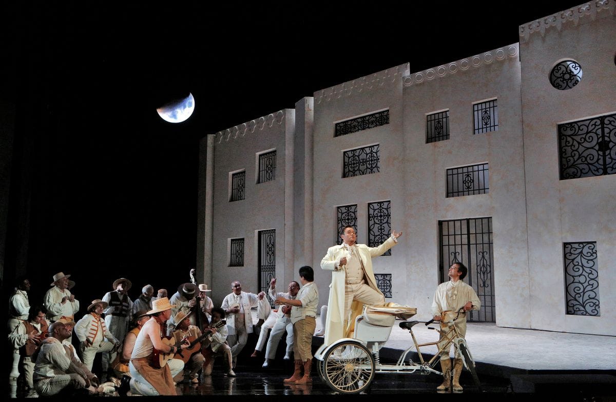 A large group of people stand together as a man stands behind a bike speaking. There is a building in the background and a crescent moon in the sky.