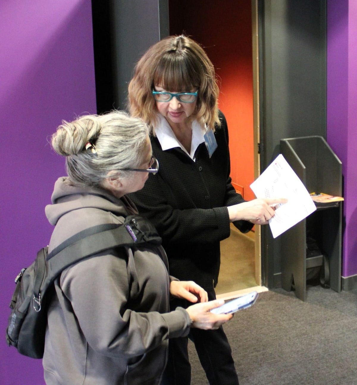 Two women stand together in a theatre lobby. One is showing the other a piece of paper and talking.