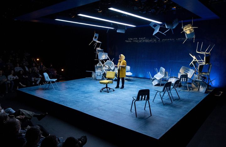 A black box theatre. A woman wearing yellow stands in front of an audience. There are chairs stacked around the stage.