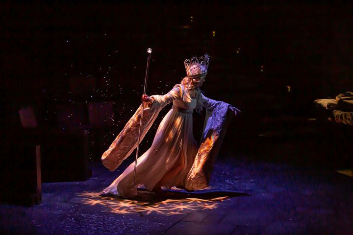 A woman wears a white gown with long, trailing sleeves. She wears an ornate crown and holds a scepter. 