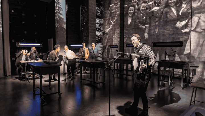 A man in the forefront plays an accordion and sings with backup singers.
