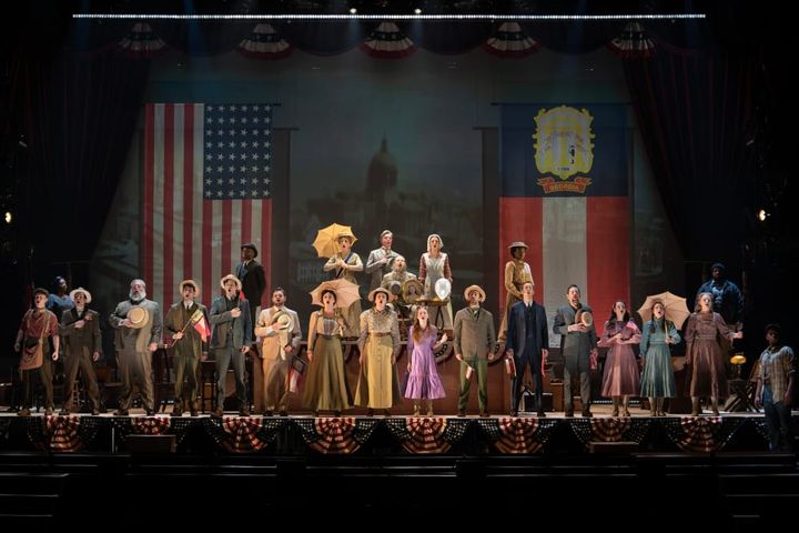 A large group of people stand in front of the American flag with hands on hearts and singing.