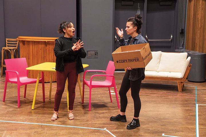Two women stand in a rehearsal room with furniture around them. They talk actively and one woman holds a cardboard box.