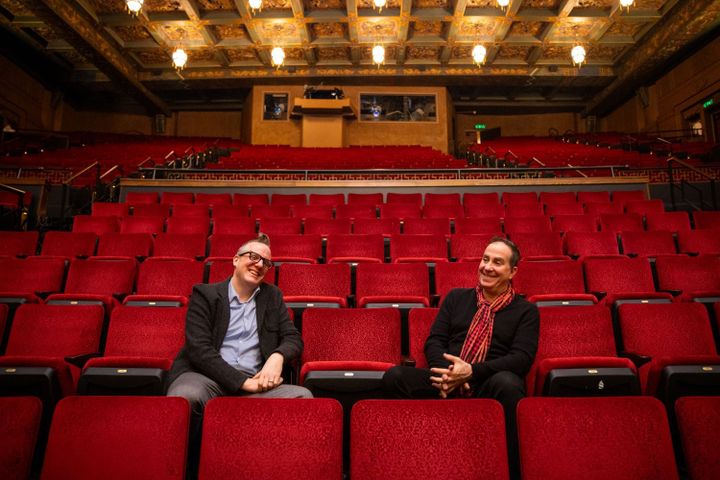 Two men sit in red audience seats in a theatre laughing.