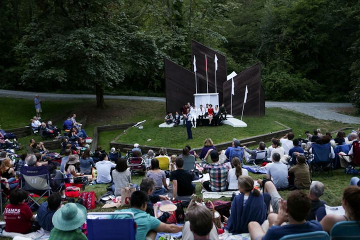 An audience of people sit outdoors in a park watching Shakespeare in the park.