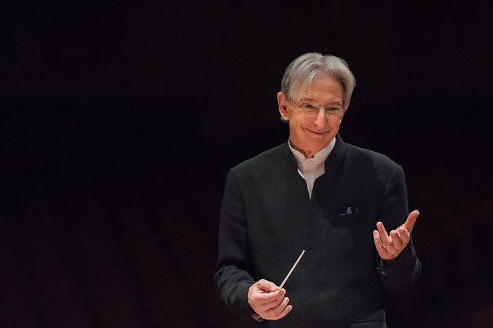 Michael Tilson Thomas stands in front of a black background smiling and holding a conductor's baton.