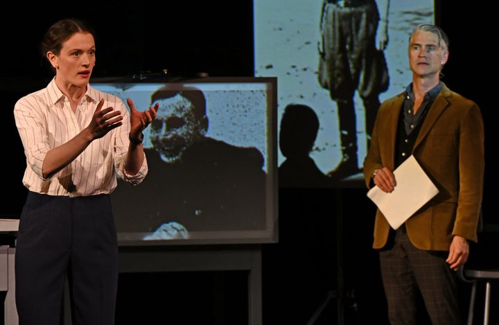A woman stands talking and gesturing with her hands while a man stand holding paper watching. Black and white photos are in the background.