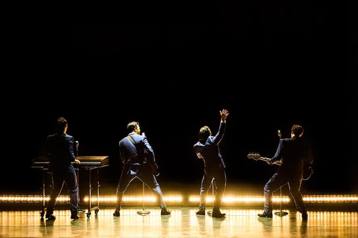 Four musicians on a stage perform with stage lights illuminating them.