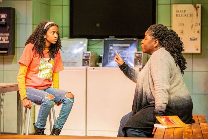 A young teen girl sits on a stool and listens as an older woman talks to her in a laundromat. 