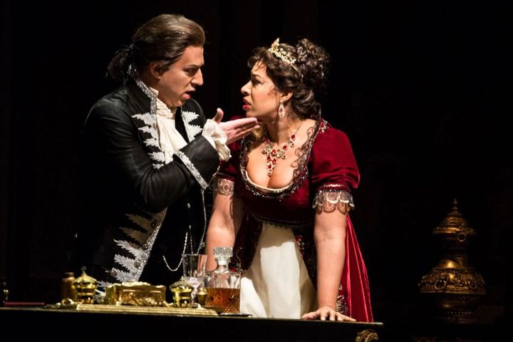 A man and woman wear 18th century clothing. The man holds his hand on the woman's chin and they look at each other intensely.