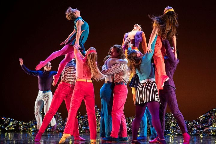 A group of dancers in colorful cloths are in a group: two hug and others hold up female dancers.