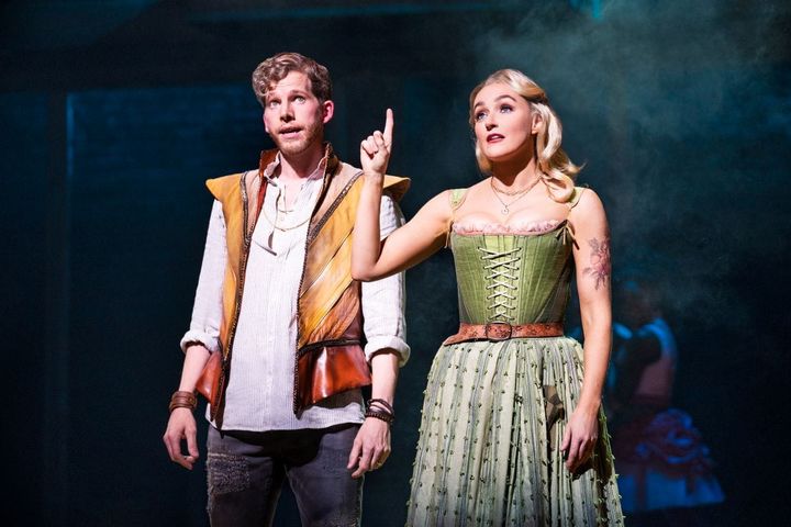 A man and woman wear modernized 16th century clothing. The woman holds her hand and pointer finger up.