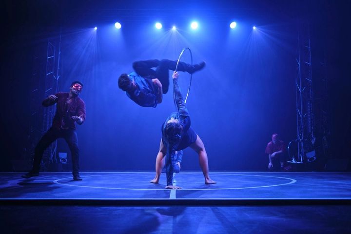 On a blue lit stage, a woman bends over backwards while holding a hoop that a man flips through.