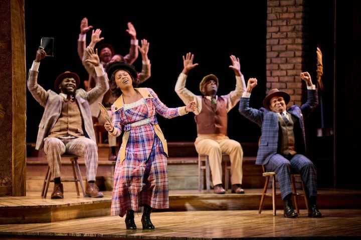 A woman sings in front of four men sitting in chairs with their hands raised. They all wear 1930s clothing.