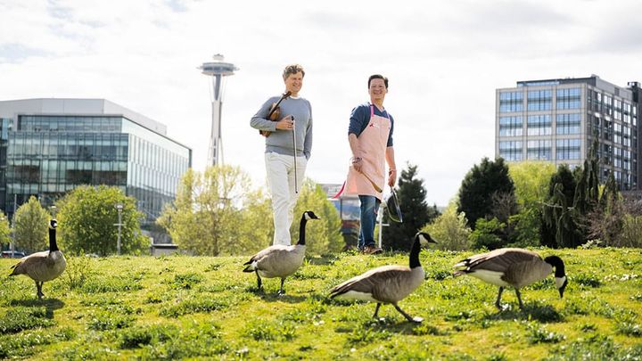 James Ehnes holds a violin and Kenji López-Alt a pan and wooden spoon on a grassy knoll with the Space Needle behind and geese in front.