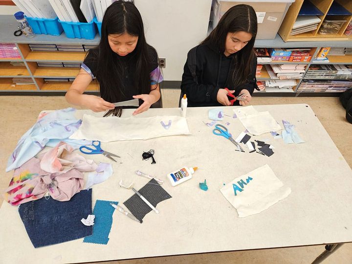 Two young girls sit at a table cutting up fabric and designing.