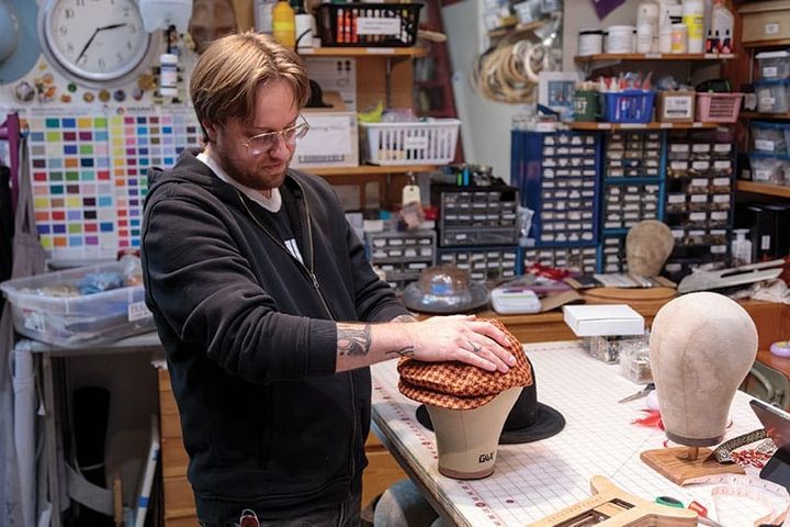 A man works in a workshop, working on a hat which rests on a head bust.