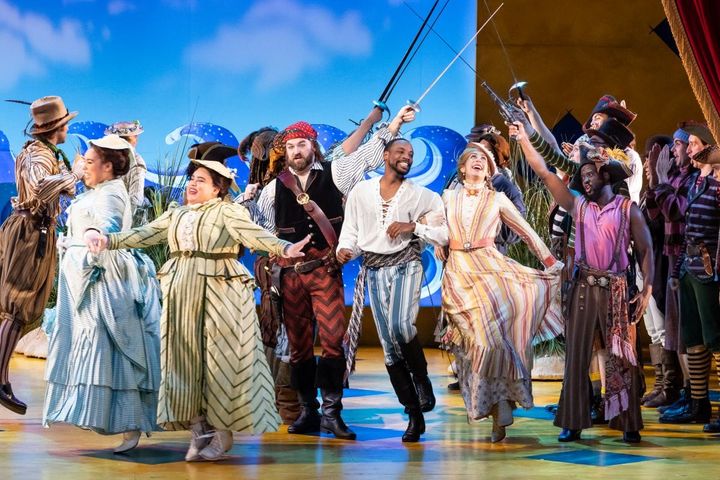 A large group of people in 19th century clothing dance around while pirates hold swords in the air.