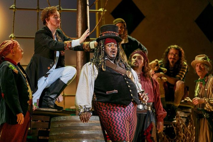 A group dress as pirates stand on a staged ship, one is front and center looking surprised.