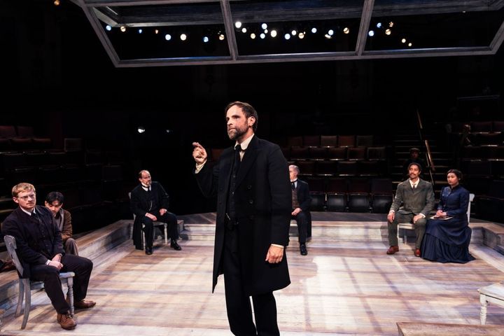 On stage a group of people sit in a semi circle looking at a man standing who is talking.