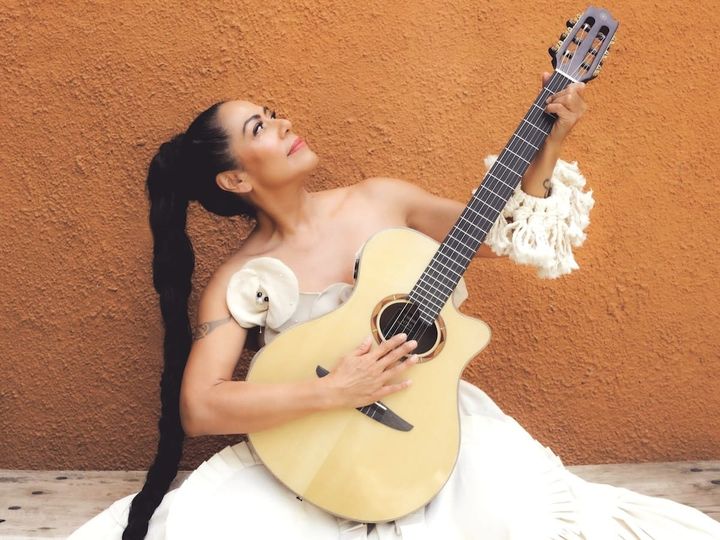 Lila downs wears a white dress, with a long braid. She sits in front of a beige wall and holds her guitar.