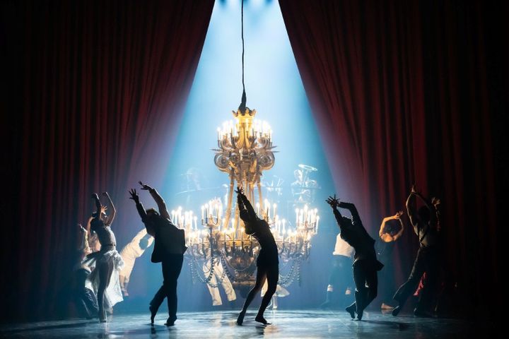 A group of dancers are on stage, some are in shadow. They dance in a circle around a chandelier that is close to the stage.