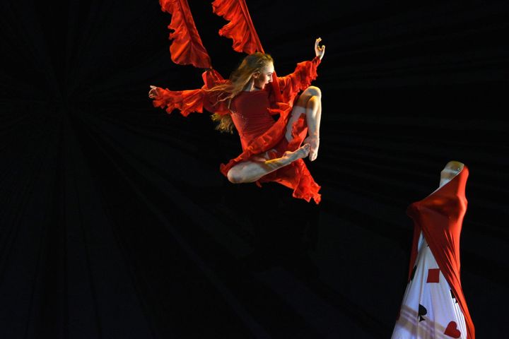 Against a completely black background, a dancer is doing an aerial stunt, flying at a woman dressed in a red and white outfit.