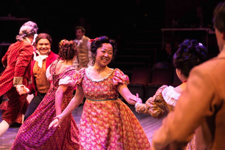 A group of people in 19th century clothing dance together in a group, smiling.