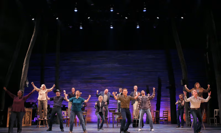 A large group of people on stage have their arms raised and jump for joy.