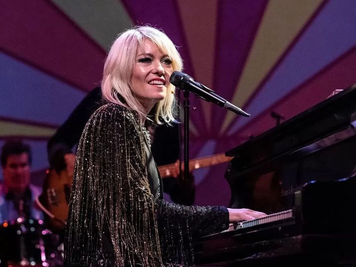 Jen Ayers looks at the audience as they are on stage playing the piano and has a microphone in front of them.