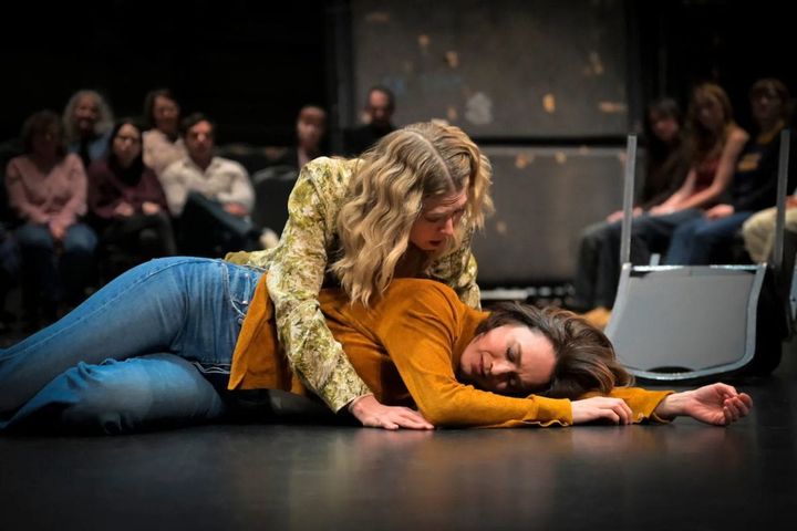 A woman is passed out on the ground, another woman leans over her looking concerned. The audience looks on in the background.
