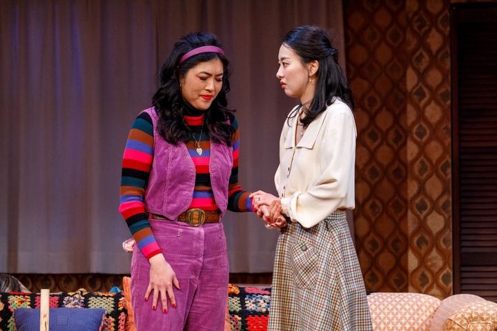 Two women stand together holding each others hands looking thoughtful.