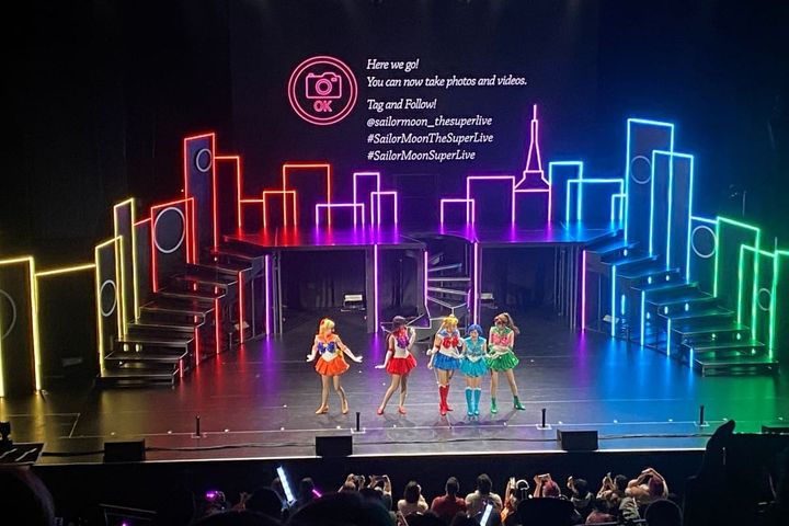 On stage a colorful light up set shows a skyline and on a screen are sing a long words. On stage are the sailor guardia