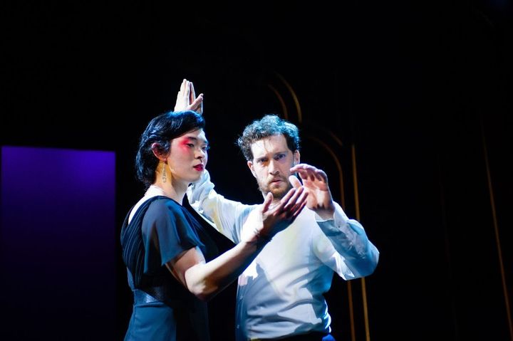 A man dressed in traditional Chinese stage makeup dances with another man wearing a suit.