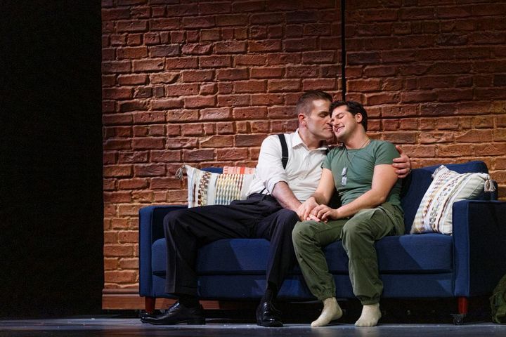 Two men sit on a coach holding each other with their heads together.