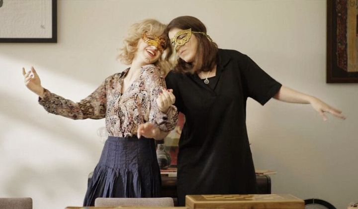 Two women lean against each other as they dance joyfully wearing masks.