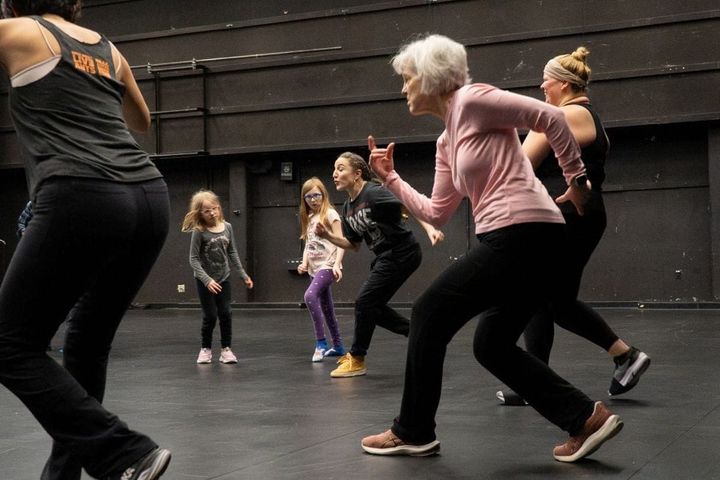 A range of women from young girls to older dance in a black box studio.