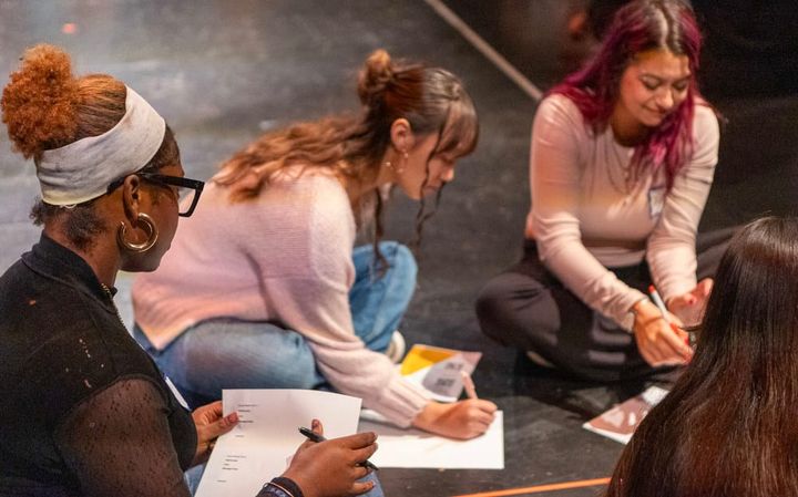 Four women sit on the floor writing and working together.