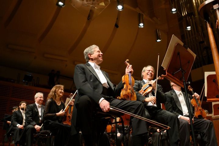 Violin players sit on stage in between playing pieces as they smile and laugh.