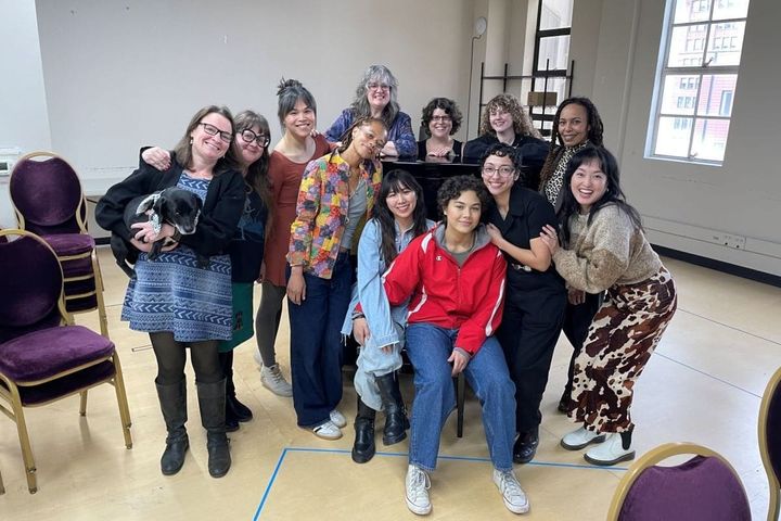 A group of women are in a rehearsal room grouped around a piano and smiling for the camera.