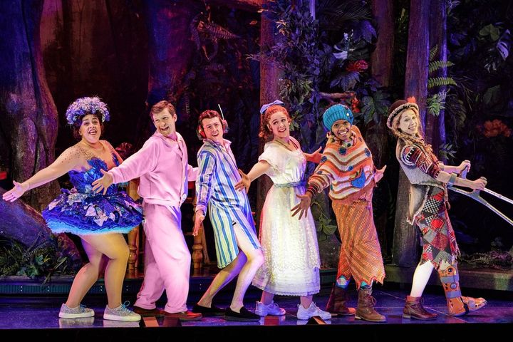Actors dressed in Peter Pan costumes are in a line in front of a forest background as they sin