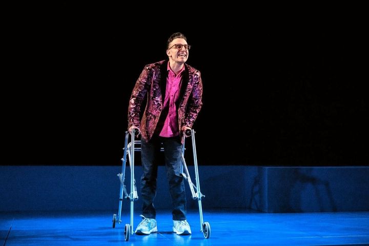 A man wearing jeans, a pink shirt and multicolored blazer stands with a walker on a blank stage cast in blue.